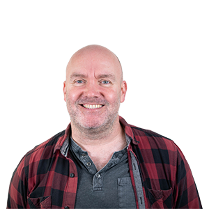 A photograph of CHAOS Radio presenter Dom Philbin; he is wearing an open red and black checked shirt with a washed-grey polo underneath.