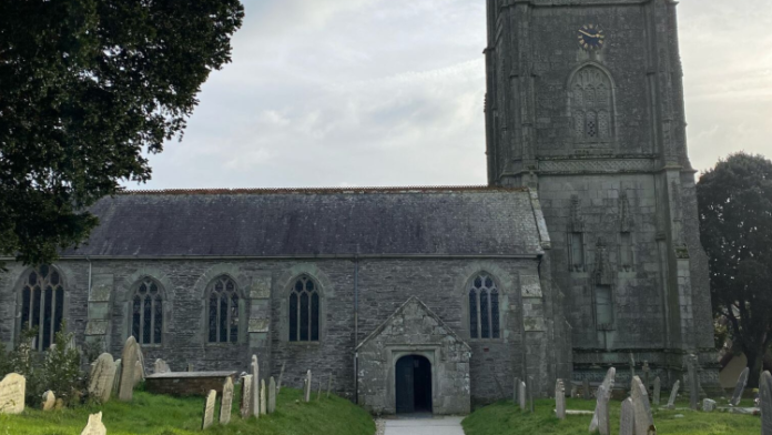 A photograph of Probus Parish Church, in Probus Cornwall. The day is sunny but overcast, with tombstones and grass on either side of a walkway that leads to the church door. A tree filled with green foliage is to the left.