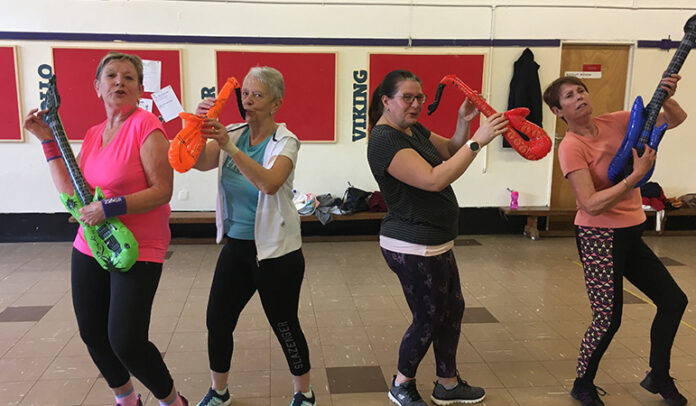 A photograph of four female members of the Zumba Club that is hosted at Lostwithiel Community Centre. They are each holding blow-up guitars and saxophones, and all wearing workout outfits and trainers - ready for Zumba.