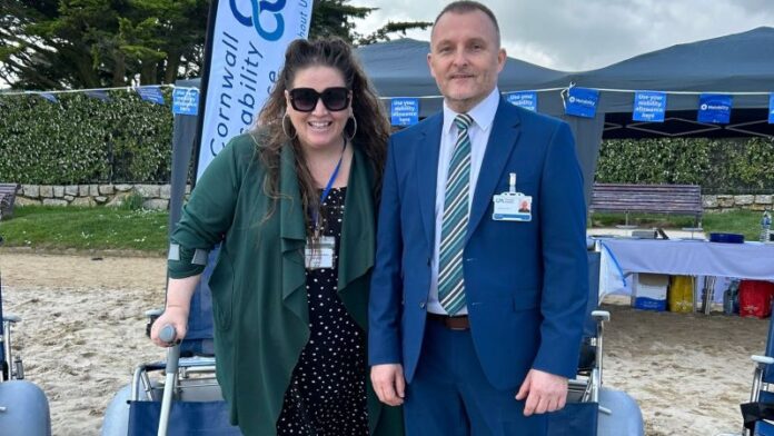 the image is of two Cornwall Mobility staff stood on the beach. Left, female, wears a green waterfall cardigan over a black and white polkadot dress, large brown sunglasses, and large gold-hooped earrings; she has long wavy brown hair and uses a crutch. Right, male wears a royal blue suit, light blue shirt, and a striped white and blue tie. Both are wearing Cornwall Mobility ID badges.