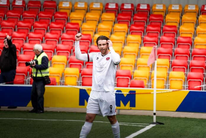 the image is a photograph of Truro City FC's Tyler, walking across the pitch with a hand up in celebration.