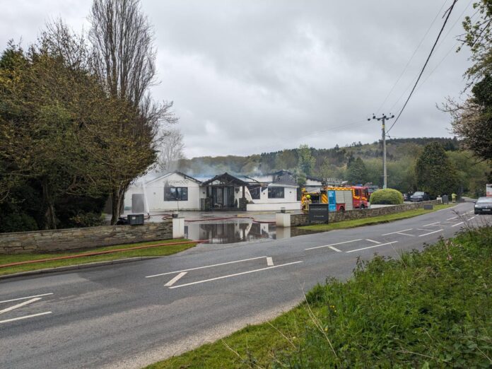 the image is a photograph of the Kingswood Restaurant, still smoking and burning, with a fire engine and staff outside.