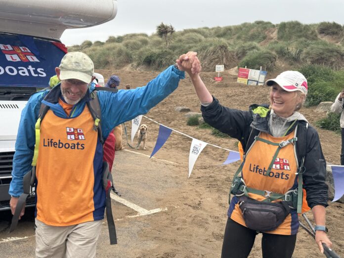 the image is of Sally and Antony Brown, walking into the finish line at a local beach in Cornwall. There is bunting behind them. They are both wearing outdoor walking gear and RNLI Lifeboat jackets.
