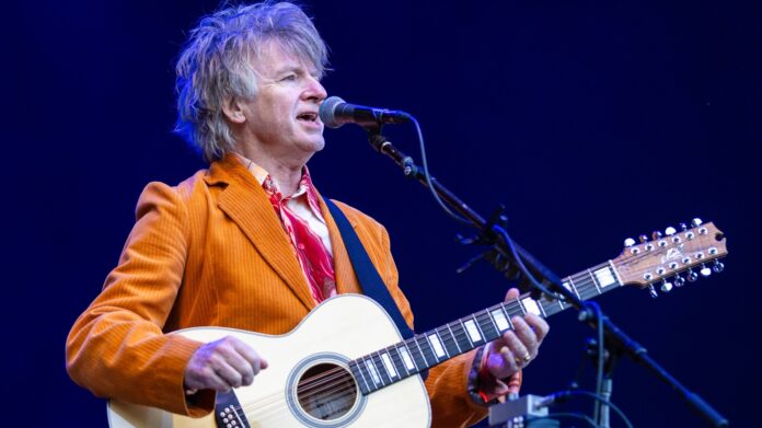 the image is of Neil Finn musician on stage at the Eden Sessions; he is wearing a bright orange corduroy jacket, deep purple trousers, a white and red shirt. He is holding a guitar and singing into a microphone.