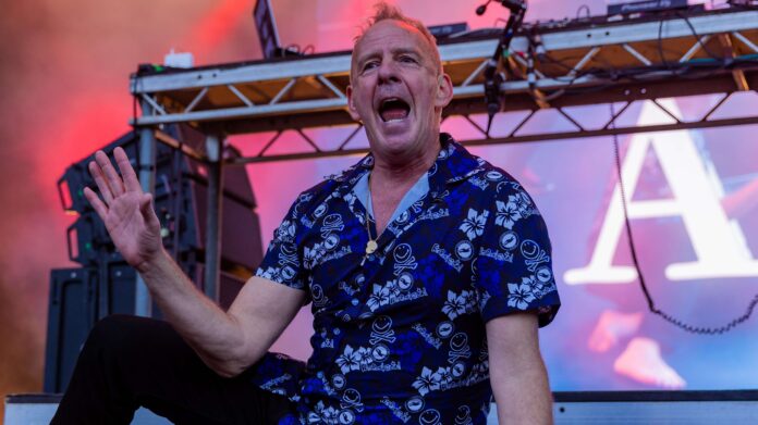 the image is a photograph of musician Fatboy Slim sat on the stage and cheering to the crowd; he is sat in front of his DJ desk and technical gear, and in front of an illuminated screen.