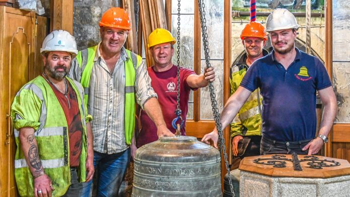 Five men (3 in high vis jackets, two in tops with bell logos on the chest), all wearing hardhats stand alongside the lowered church bells.