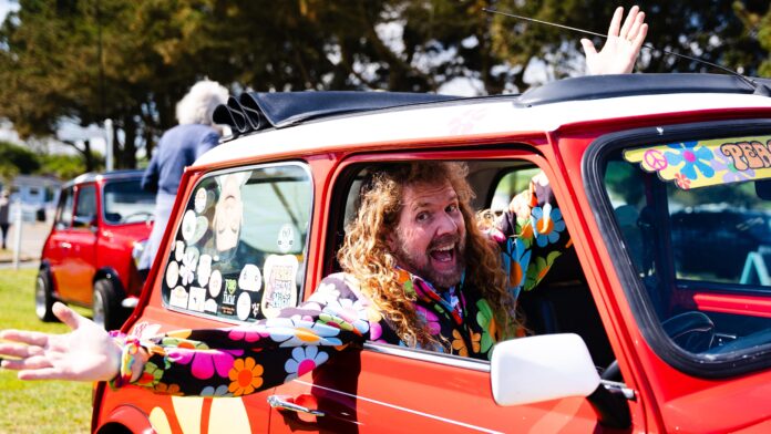 Mike Guido sits inside his red mini, covered in stickers. He wears a black jacket covered in very vibrant flowers.