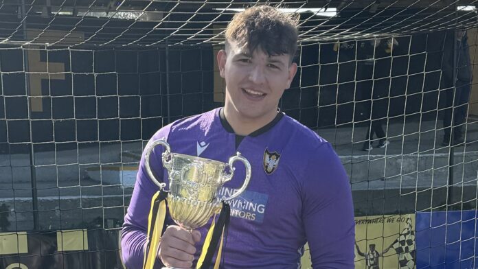 Morgan Coxhead (footballer) stands in front of a goal, holding a trophy. He wears a purple football shirt for Falmouth Town FC.