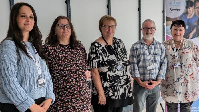 Seetec Pluss Employability Support Day; five people stand in a line next to each other, all wearing printed and patterned clothes. At the end of their line is a large marketing board from Seetec Pluss, that mentions wording in line with the event.