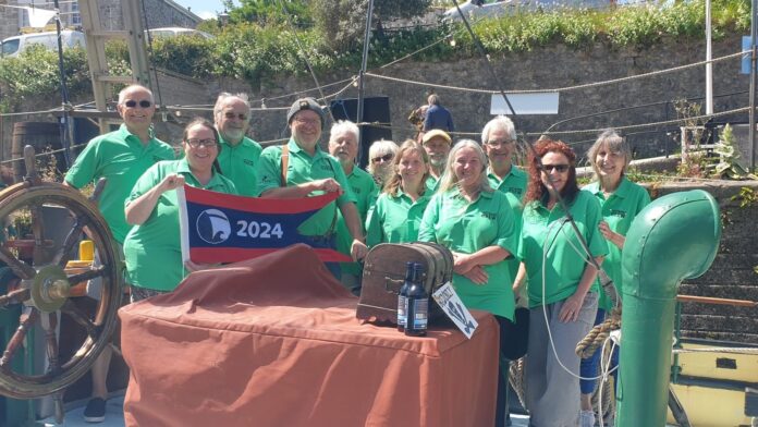 CHAOS Radio Newswave - a group of 12 volunteers stood aboard the old ship 'The Lady Daphne' within Charlestown, Cornwall - they all wear matching green polo shirts with a black embroidered 'CREW' emblem on the left chest. To the left, two volunteers hold a red and navy flag that reads '2024'. In the centre is a table covered in a red cloth, with a wooden pirate chest to the right.