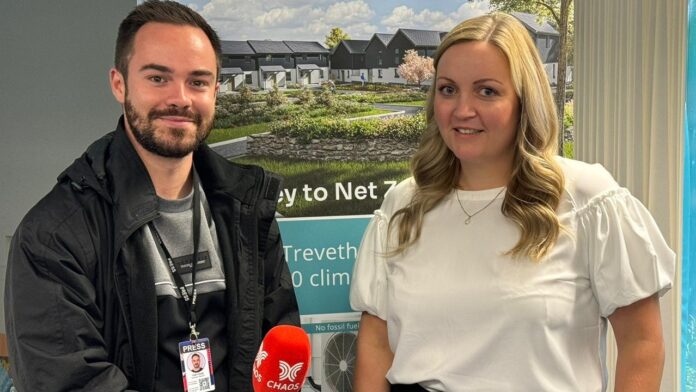 CHAOS Radio Newswave - CHAOS Radio Journalist (left) wears a black and grey jumper beneath a black coat, with a black 'PRESS' lanyard, and holds a black CHAOS microphone with red muff that has the white CHAOS Digital logo. Right is Emma Rodgers wearing a white puff-sleeve blouse and silver pendant necklace. They stand in front of a roll-down banner that features images of a housing estate.