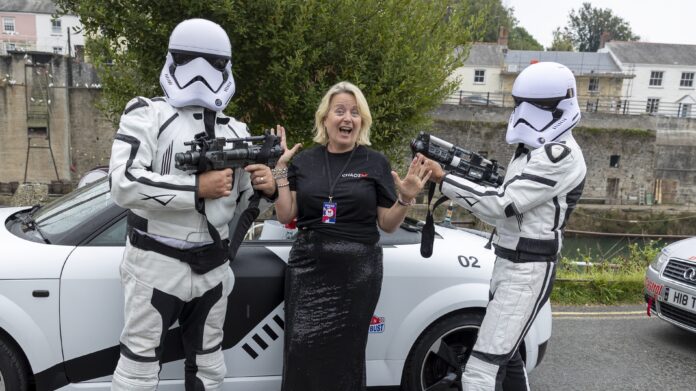 CHAOS Radio Newswave - Presenter Janice Rose is stood at Charlestown harbour, in front of a white car. She is wearing a long black sequin skirt and a black tshirt with the white CHAOS logo and red butterfly on the left chest. She is stood between two men dressed as Star Wars storm troopers, holding guns toward her.