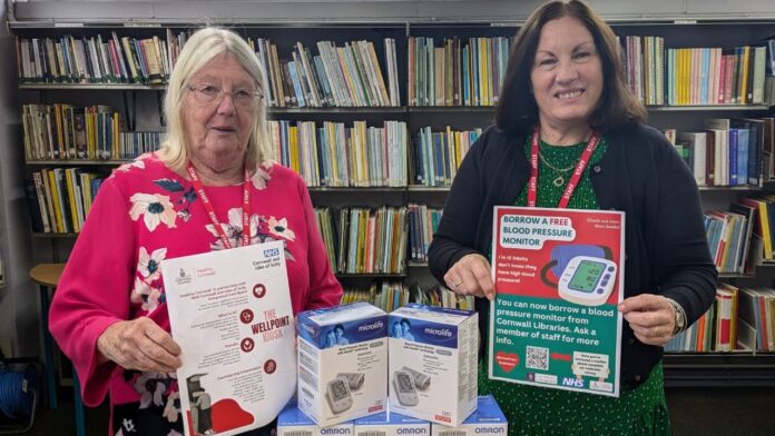 CHAOS Radio Newswave - two women stand facing the camera, each holding A4 printed sheets that promote the borrowing of a free blood pressure monitor from your local library. They stand in a library, in front of large rows of books. On the table between them are OMRON blood pressure monitors in boxes.