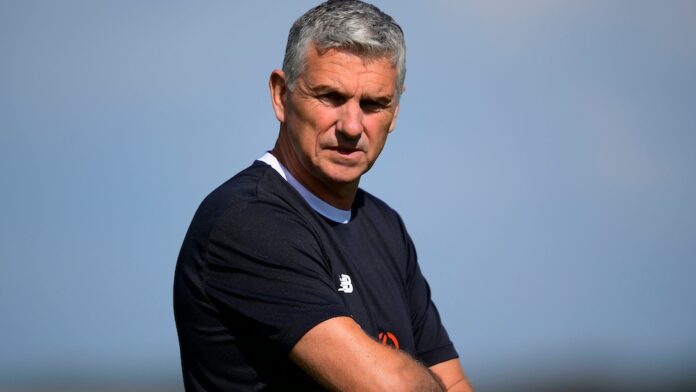 CHAOS Radio Newswave - Truro FC's John Askey stands with his arms folded, wearing a black tshirt.